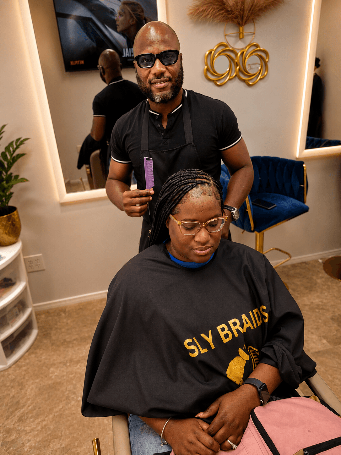 Stylist Sly working on client wearing SLY BRAIDS cape — salon interior